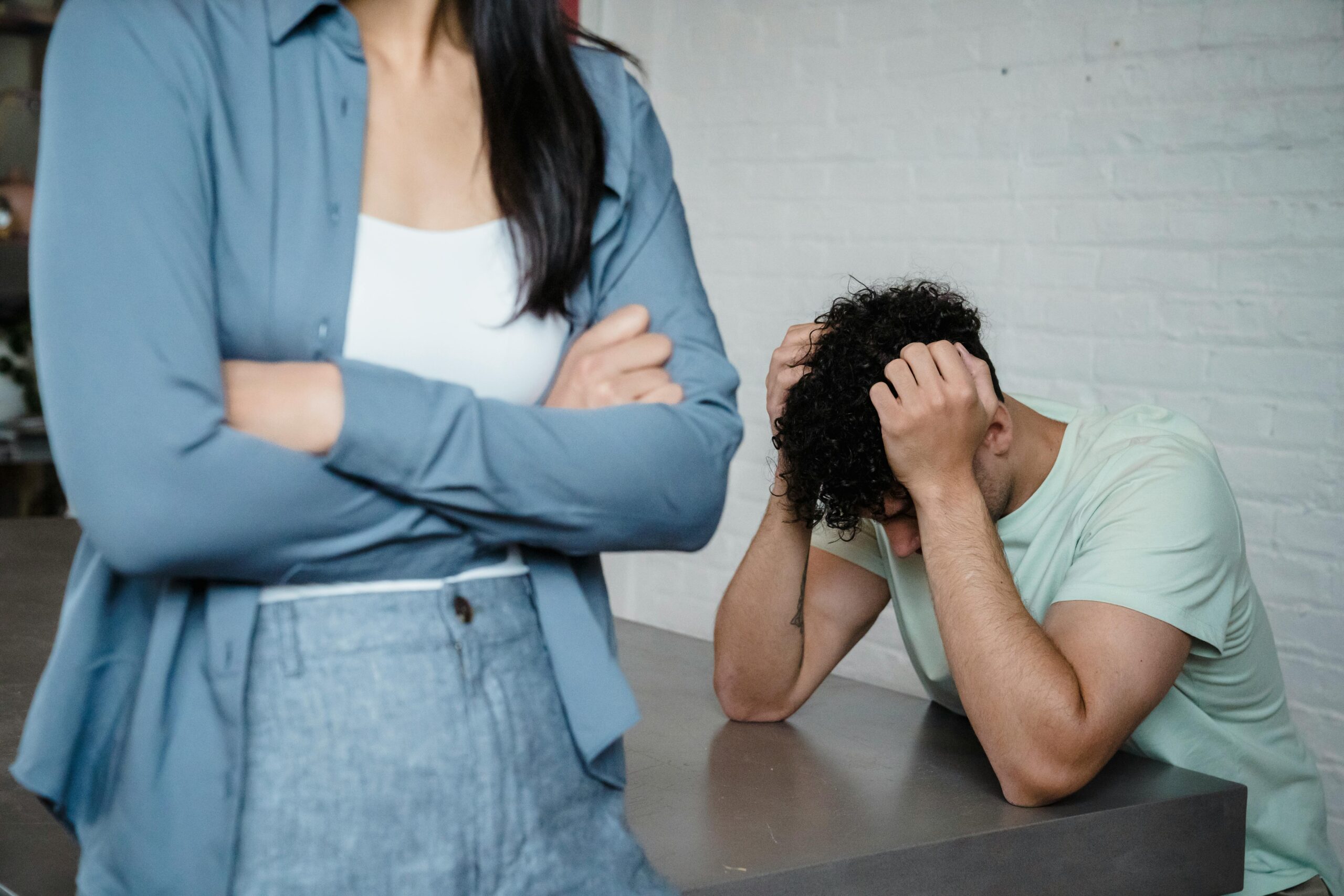 An image of a couple arguing in their living room, showcasing relationship conflict. The man sits with his head in his hands, expressing distress and frustration, while the woman stands a few feet away with crossed arms and an upset expression. The distance between them highlights emotional disconnection, emphasizing the challenges of effective communication in relationships.